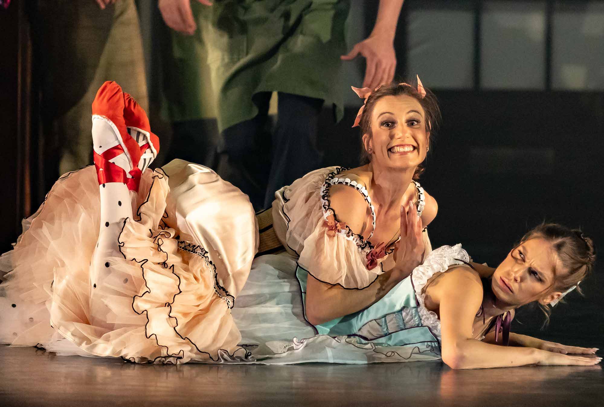 Two women in colorful, frilly dresses lie on the floor during a stage performance; one smiles widely with clasped hands, while the other looks to the side with a puzzled expression.